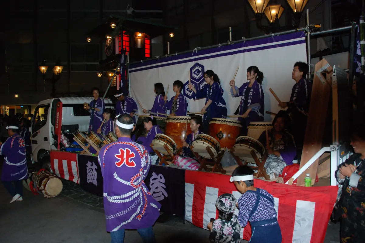 Drummers in purple festival coats play a row of taiko on a decorated float at night, under red-and-white curtains