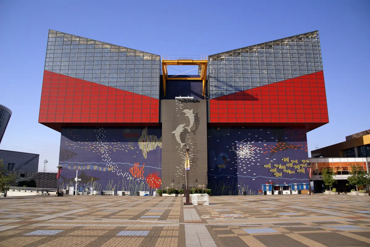 The red and blue-grey front of the Osaka Aquarium Kaiyukan seen across a paved plaza under a blue sky