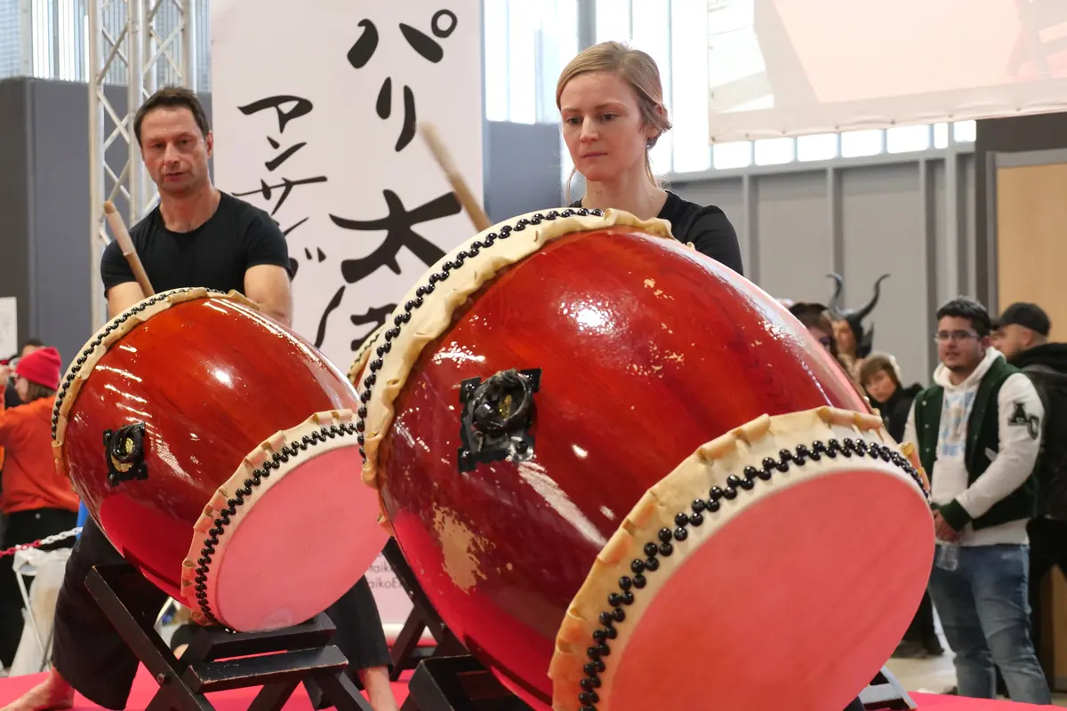 Two red taiko with cream skins and rows of studs, angled on stands, with two drummers behind them