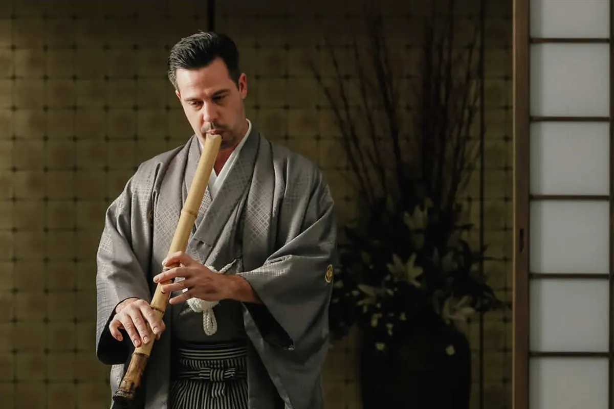 A man in a grey kimono and hakama plays a bamboo shakuhachi flute, held straight down in front of him