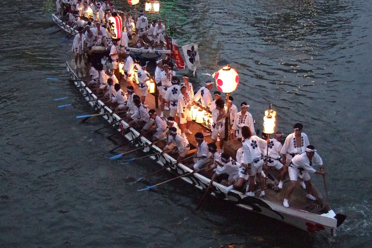 A long wooden festival boat crowded with people in white costumes and paper lanterns moves across dark water